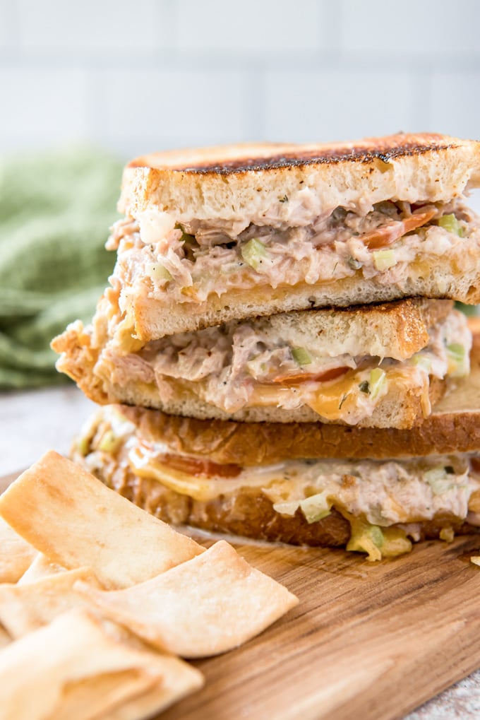 3 tuna melt sandwiches stacked on top of each other, a wood cutting board, a green napkin and chips.