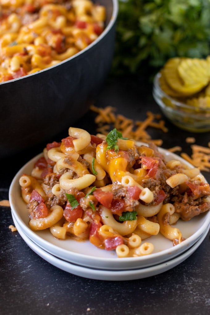 cheeseburger casserole on a white plate, a small bowl of sliced pickles, shredded cheese, a skillet with pasta casserole