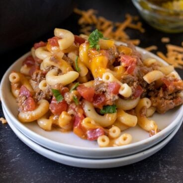 white plate stacked with cheeseburger casserole on it. Shredded cheese, a bowl of sliced pickles