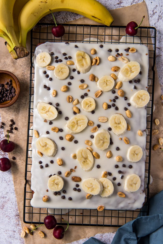frosted cake with banana slices, peanuts and chocolate chips, bananas, cherries, black wire rack and brown paper