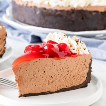 A piece of chocolate cheesecake cake on a plate, with cherry topping and whipped cream.