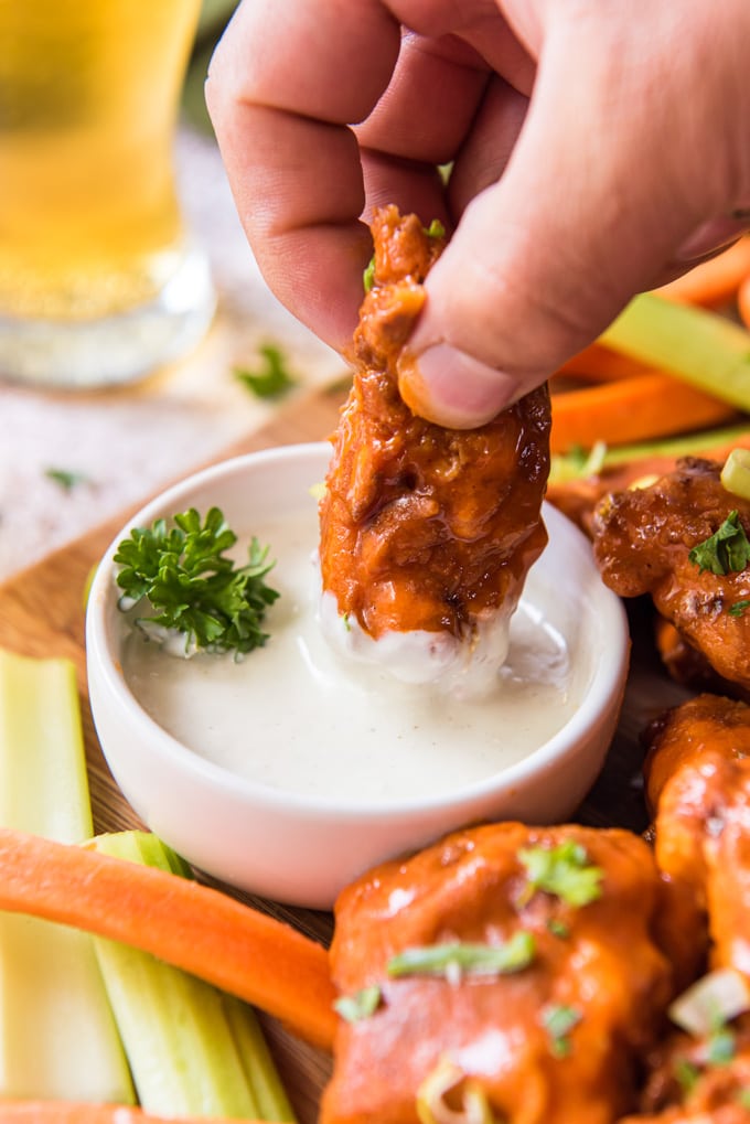 a hand holding a boneless buffalo chicken wings dipped in ranch dressing. carrots and celery sticks, a wood background, a glass of beer