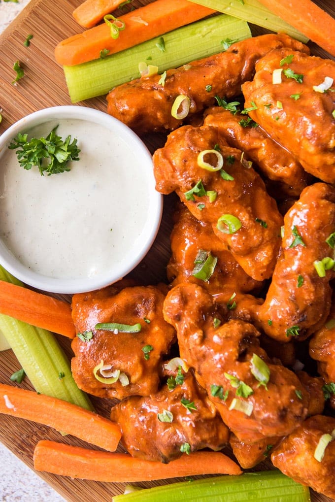 fried chicken pieces with buffalo sauce, sliced green onions, carrot and celery sticks, a white dish with ranch dressing, a wood cutting board