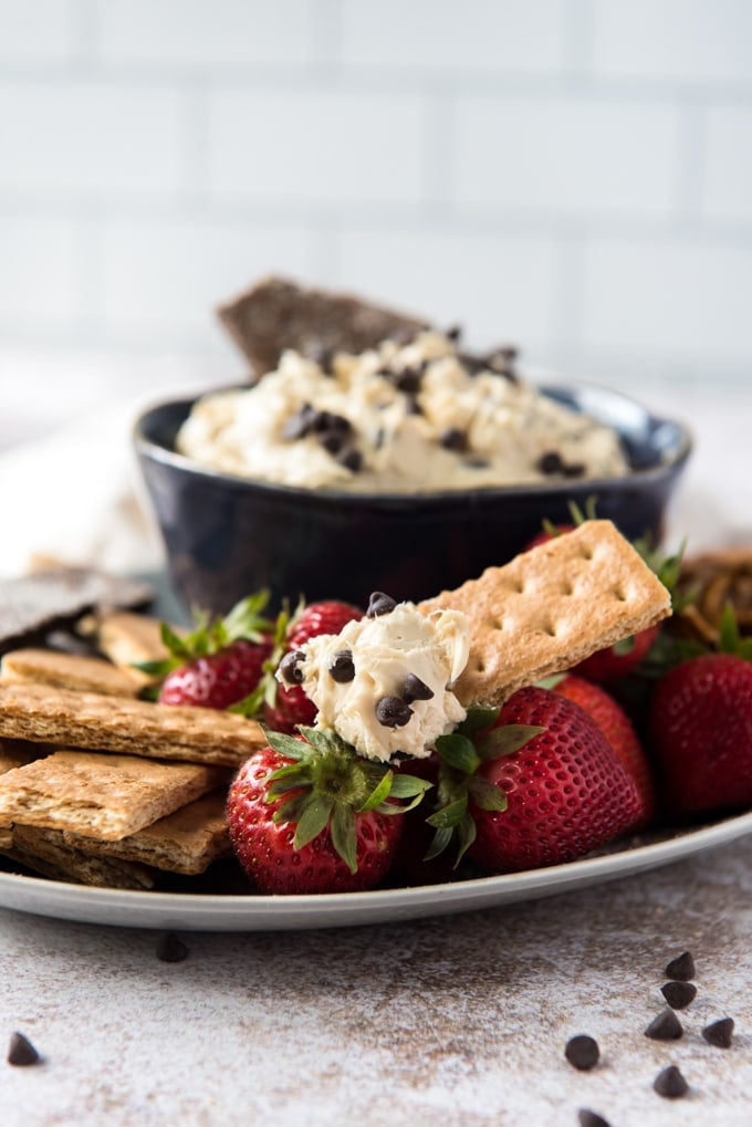 a white plate, scattered mini chocolate chips, graham crackers, strawberries cookie dough dip in a blue bowl.