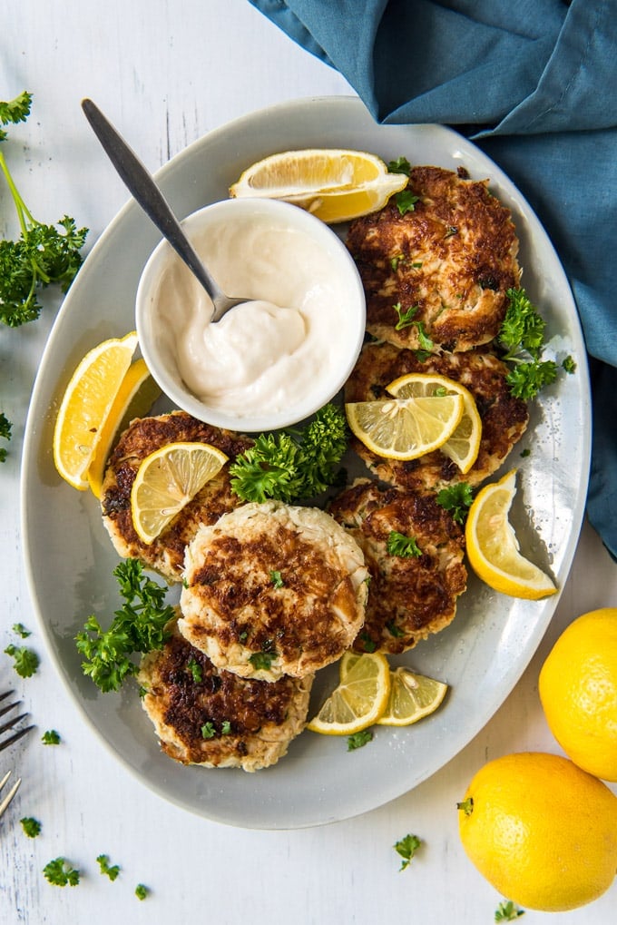 a large white platter, crab cakes, lemon slices, a bowl with tartar sauce and a spoon, a blue napkin, parsley