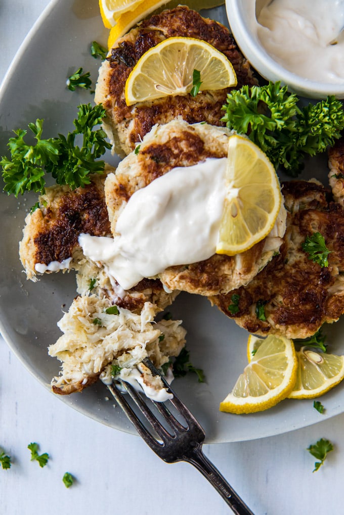 a white plate, a stack of crab cakes, lemons, tartar sauce, a crab cake pulled apart to show the interior, parsley