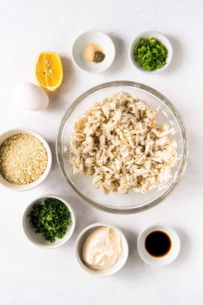 7 small white bowls with ingredients for making crab cakes. An egg, a lemon and a large clear glass bowl full of crab meat.