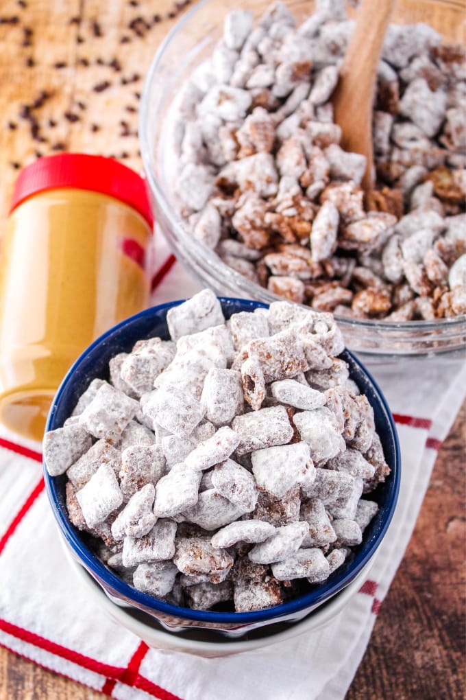 muddy buddies in a bowl with a jar of peanut butter and chocolate chips around it