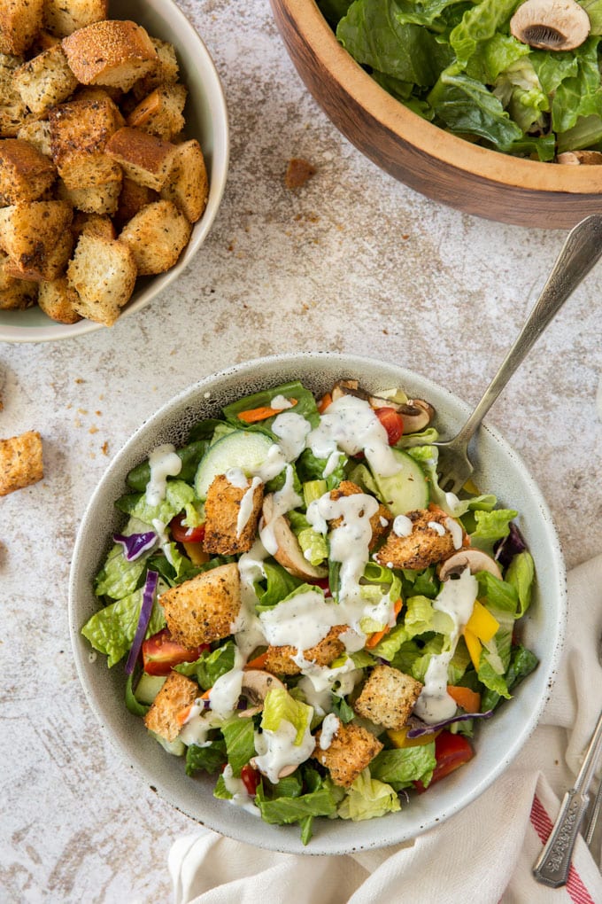 a white bow with salad, ranch dressing and a fork, a white bowl with croutons, a wood bowl with salad