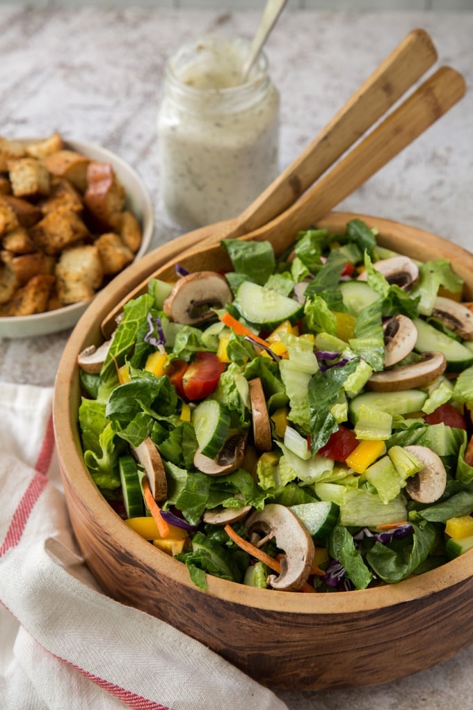 a wood bowl with fresh lettuce and other vegetables, two wood spoons, a bowl of croutons and a jar with ranch dressing and a spoon.
