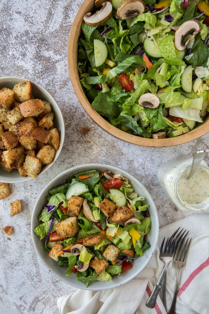 a white bowl with salad, a white bowl with croutons, forks, a white towel with a red stripe and a wooden bowl with salad and veggies, a mason jar with ranch dressing and a spoon.