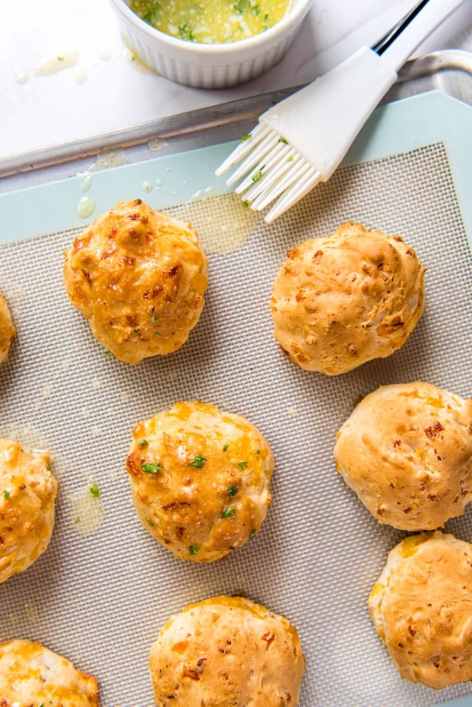 a baking sheet and a silicone mat, biscuits brushed with butter and topped with parsley, a white silicone pastry brush, a white dish with melted garlic butter.