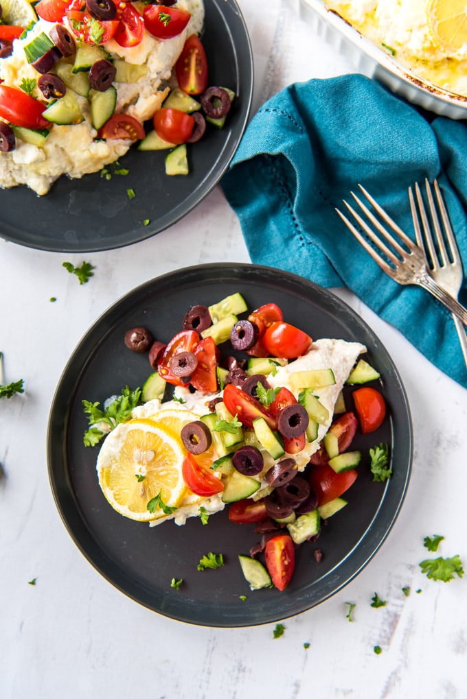 baked chicken breasts with a cheese mixture, lemon slices, olives, cucumbers, tomatoes, a blue napkin, 2 forks, two black plates