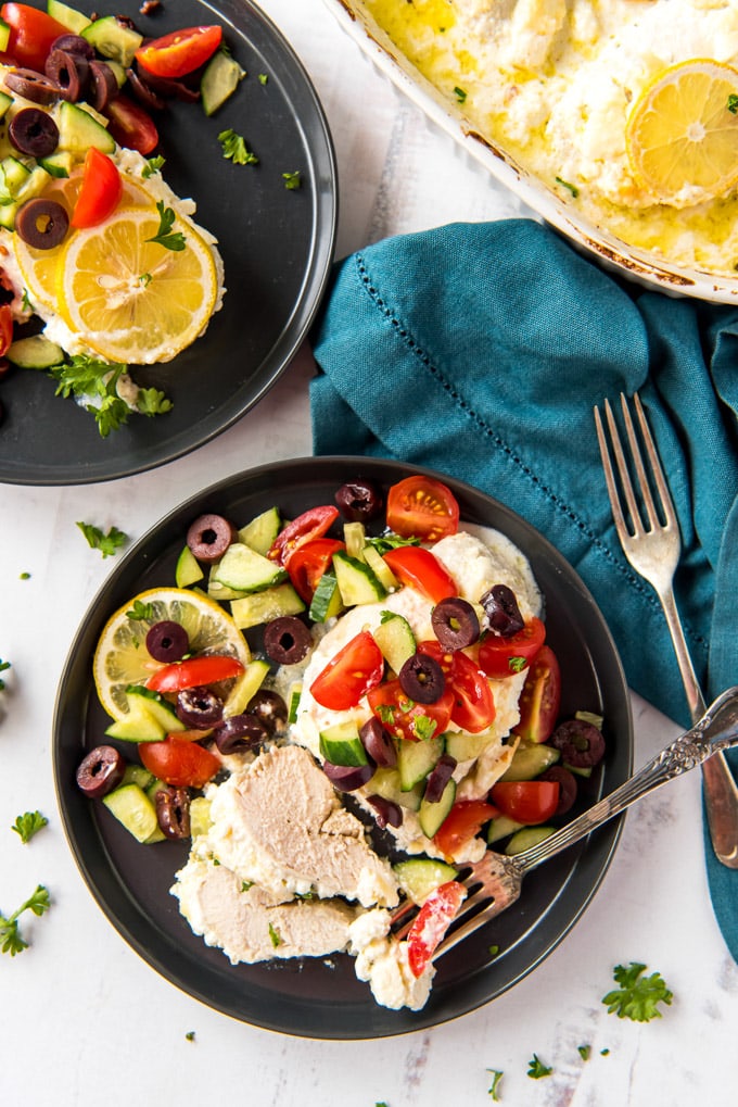 two black plates with chicken, cucumbers, olives, tomatoes, a blue napkin, a fork a whilte casserole dish with chicken and cheese in it.