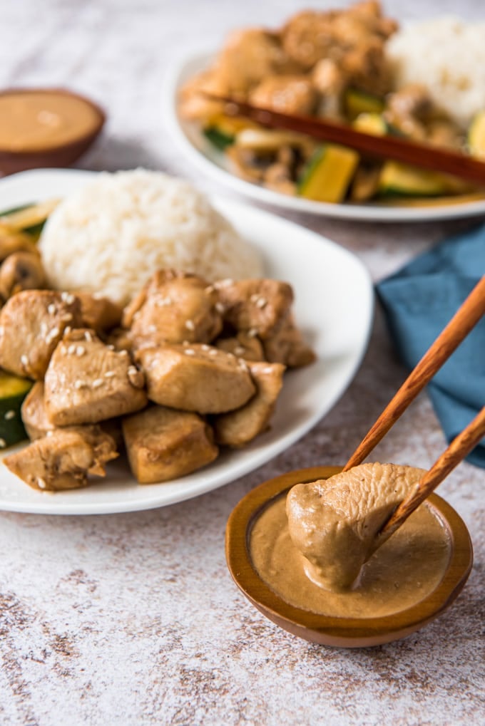 a wood bowl with yellow sauce, chicken held by chopsticks, white plates with chicken, vegetables and rice.