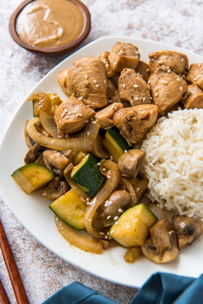 white plate with chicken, vegetables and rice, chopsticks, a wood bowl with yellow sauce, a blue napkin.