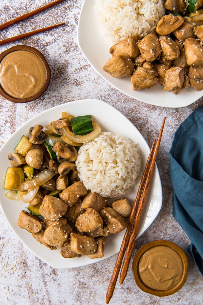 2 white plates with chicken, vegetables and rice and chopsticks. 2 small wood bowls with yellow sauce, a blue napkin.