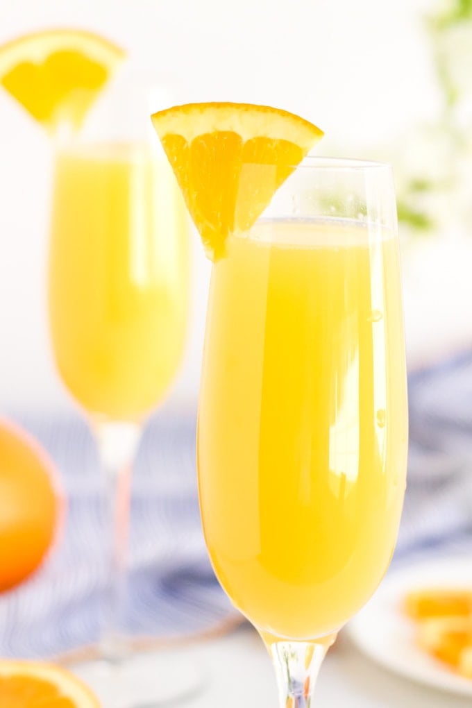 Champagne flutes with orange juice and an orange slice on the rim. A blue napkin and oranges in the background.