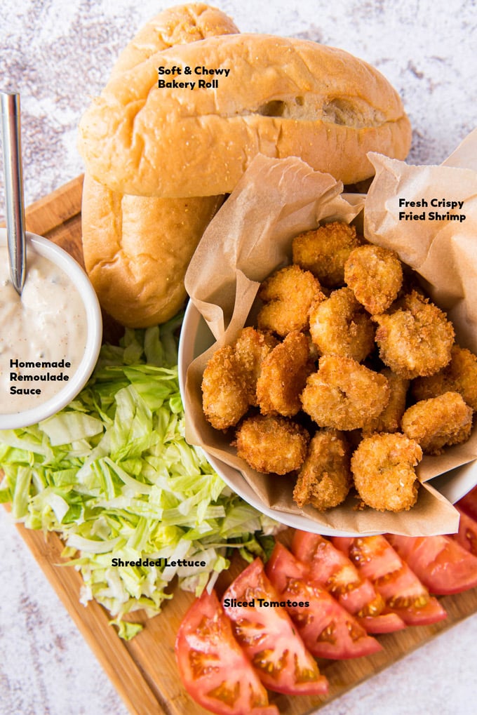 Wooden cutting board filled with hoagie rolls, fried shrimp, tomatoes, lettuce, and dressing.