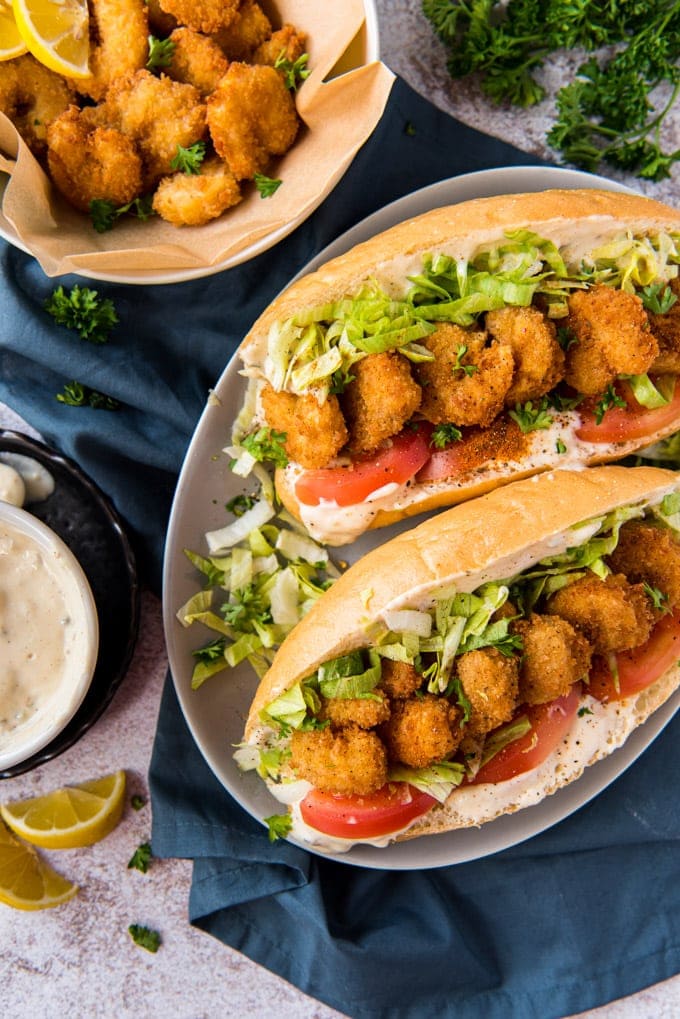 blue napkin, lemon slices, white platter, two rolls with shrimp, lettuce, tomatoes and sauce, bowl of fried shrimp.