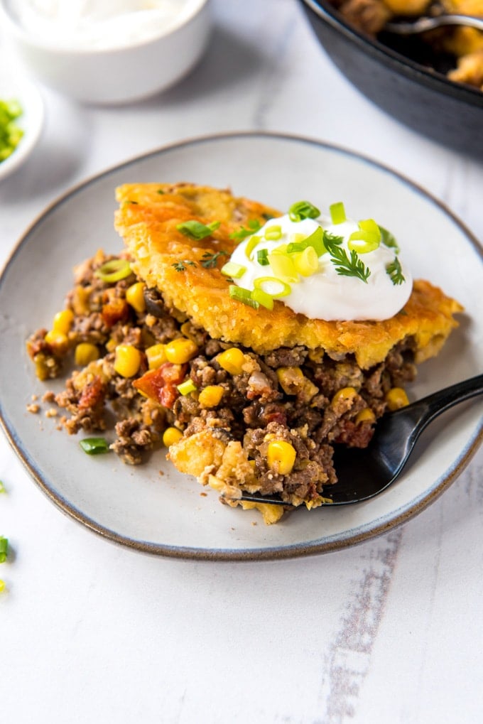 white plate with ground beef anc corn, cornbread topping and sour crea. A fork, a white dish