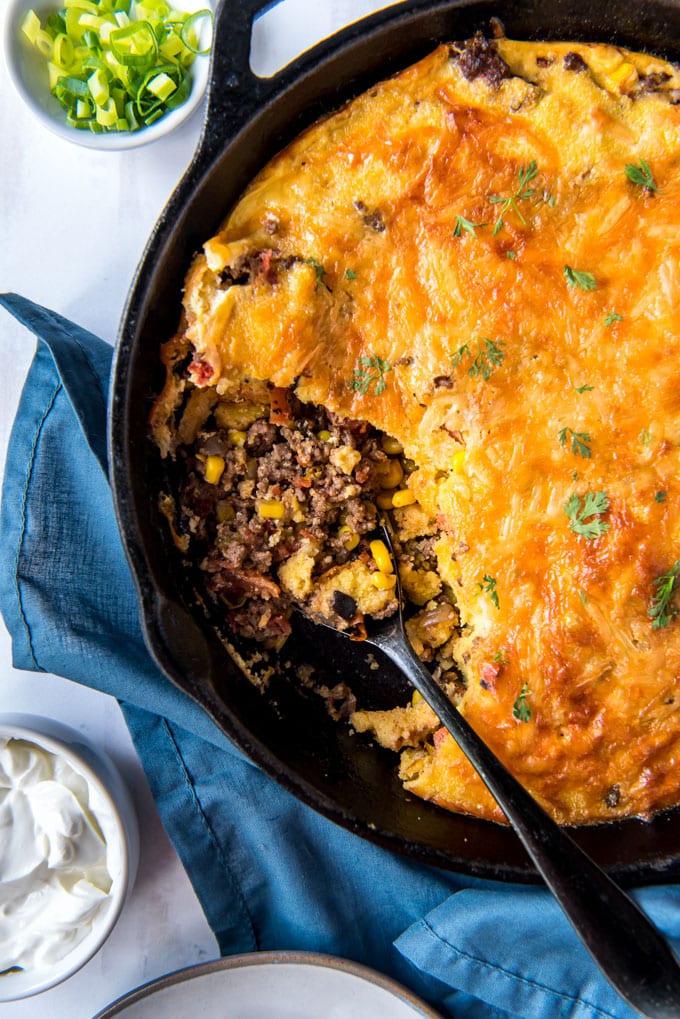 cast iron skillet with tamale pie, a spoon with ground beef with corn and tomatoes, a bowl of sour cream and a bowl of green onions