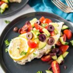 A plate with Greek chicken topped with tomatoes, olives, cucumbers, and lemon.