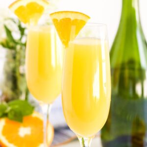 square image of 2 champagne glasses with orange juice mimosas and a green champagn bottle, an orange slice and some mint.