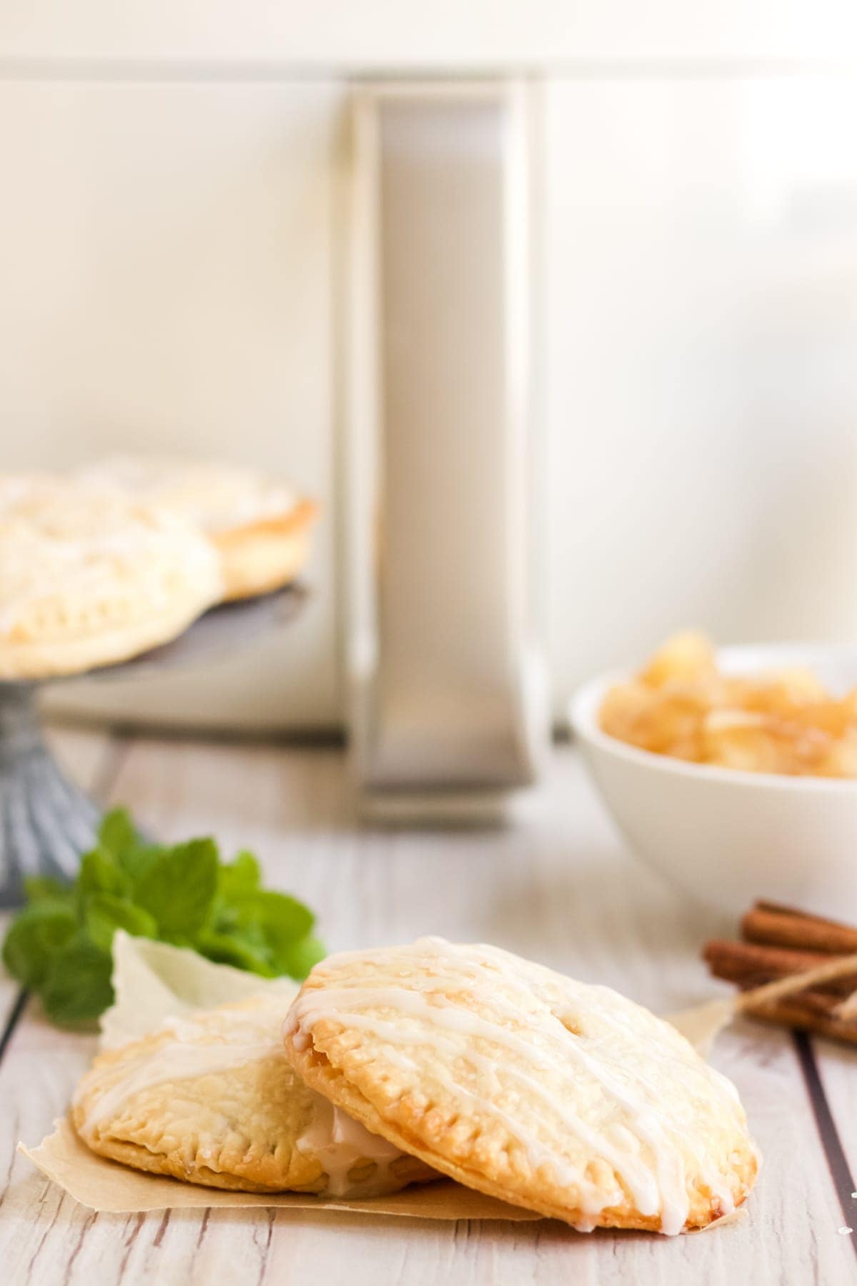 Two air fryer apple hand pies in front of the air fryer with a drizzle of glaze on top.