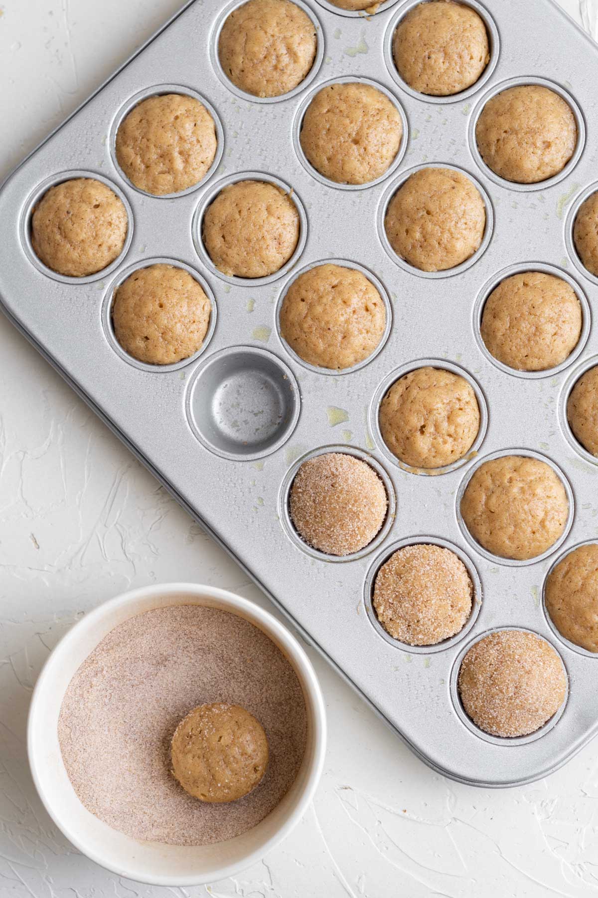 apple cider donut holes in a mini muffin pan, one sits in a white dish of sugar and cinnamon