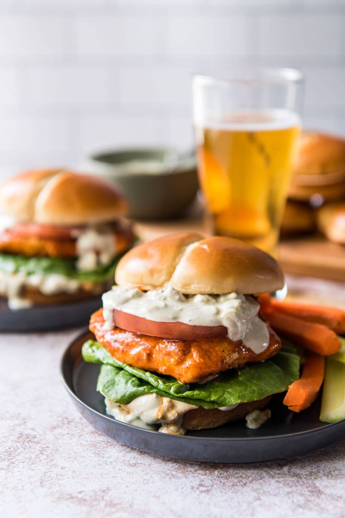 two black plates, 2 buffalo chicken sandwiches on buns with creamy sauce, lettuce and tomato, a beer, carrot and celery sticks