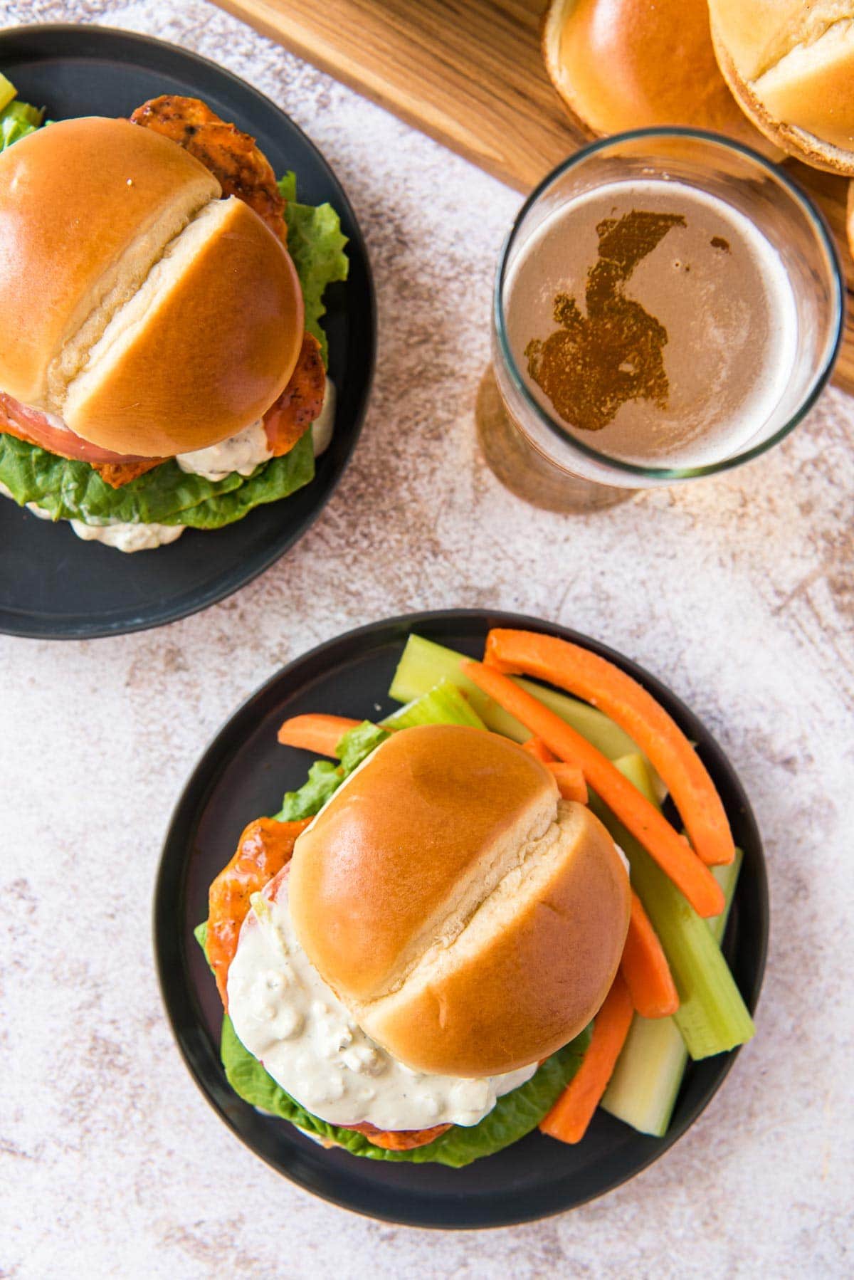 Overhead shot of 2 buffalo chicken sandwiches on black plates, a beer, brioche buns on a wood cutting board