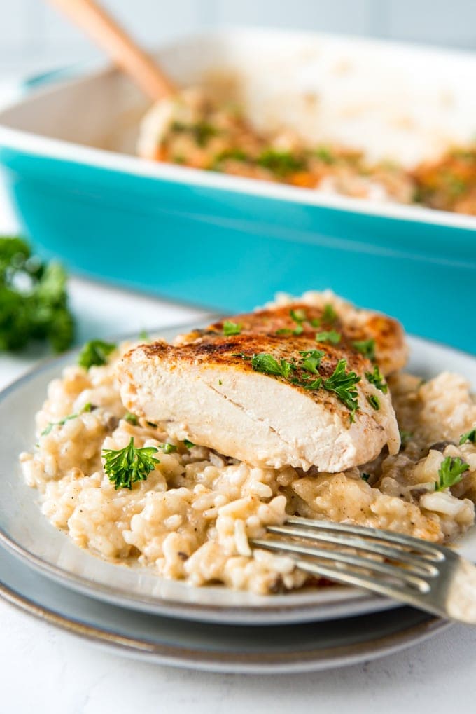 a white plate, creamy rice and half of a chicken breast, the tines of a fork, a blue casserole dish with a wooden spoon.