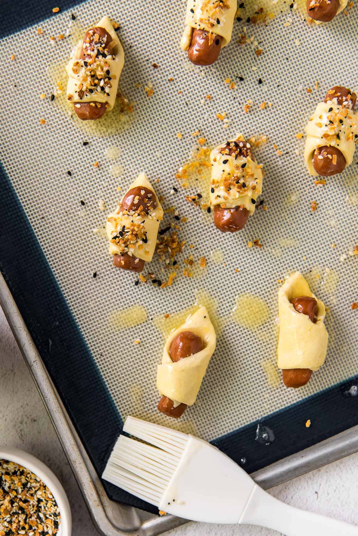 a baking sheet, white pastry brush, mini sausages wrapped in dough with bagel seasoning