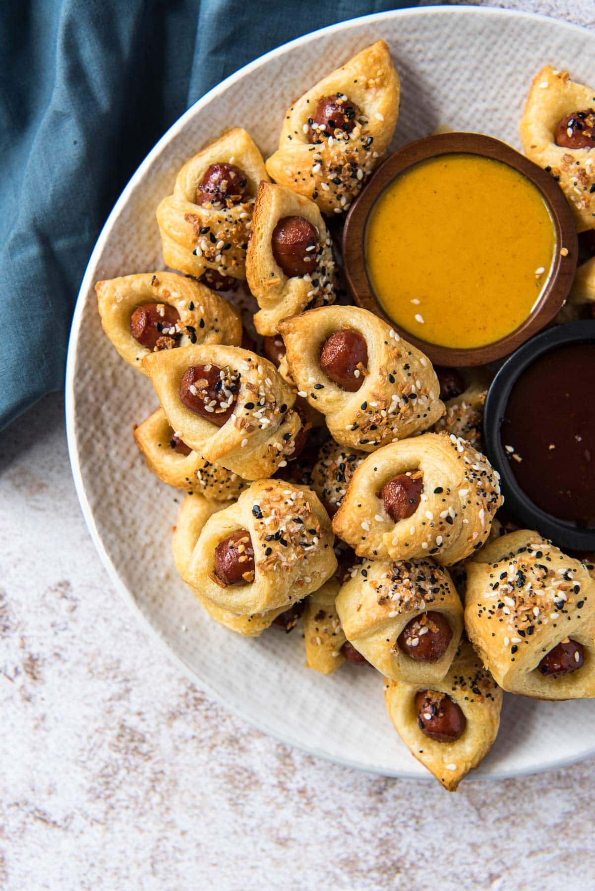 white platter with sausages wrapped in dough with bagel seasoning on top, a dish of honey mustard dip