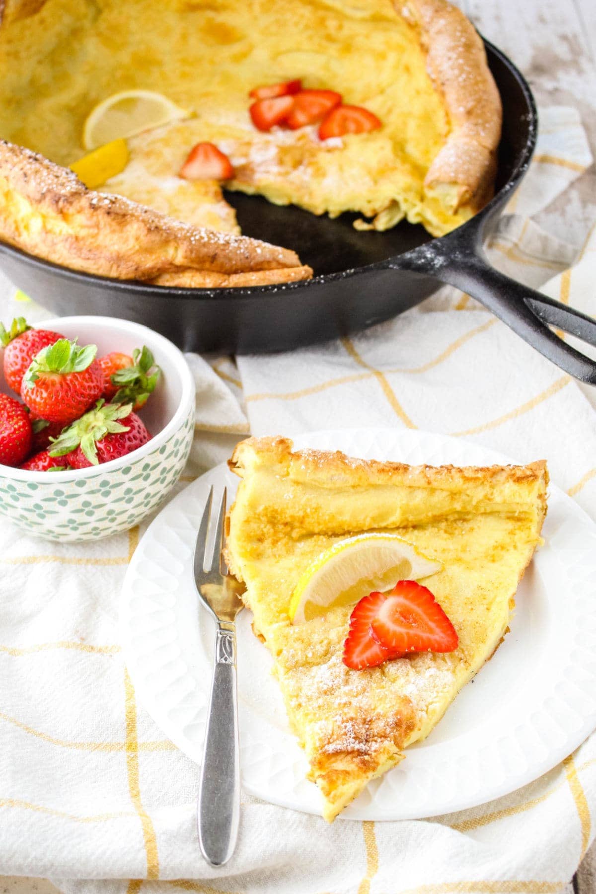 slice of german pancake on a plate with a fork next to a bowl of strawberries