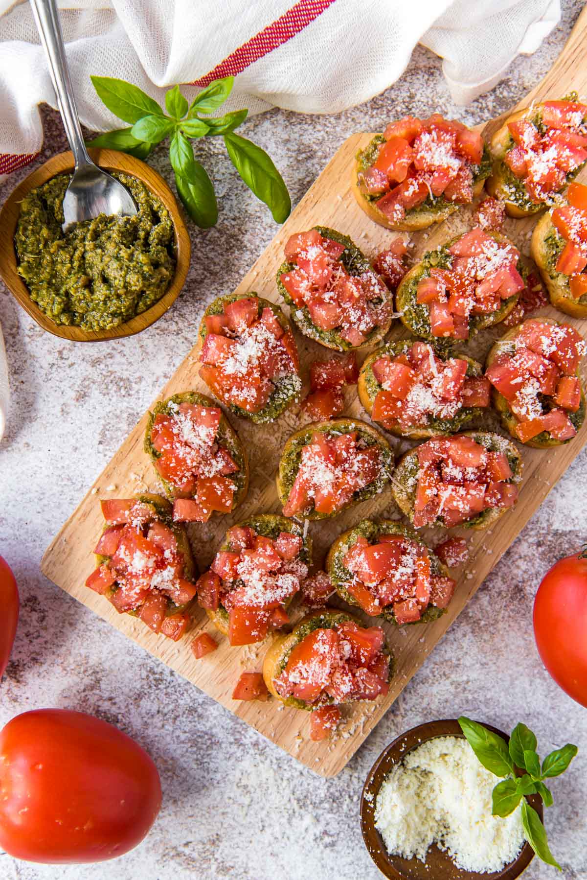 wood cutting board with bruschetta pieces, a dish of pesto, a dish of parmesan cheese, tomatoes