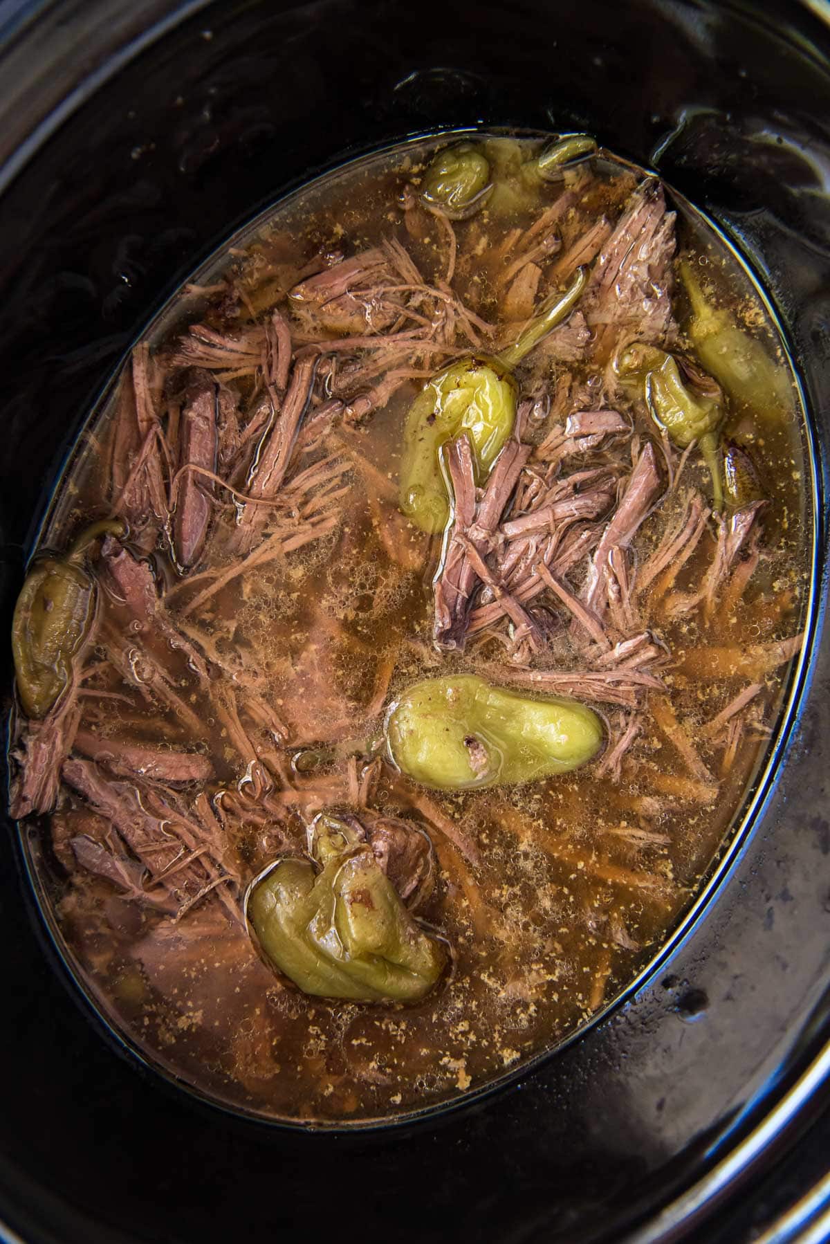 black slow cooker with shredded beef, broth and pepperoncinis