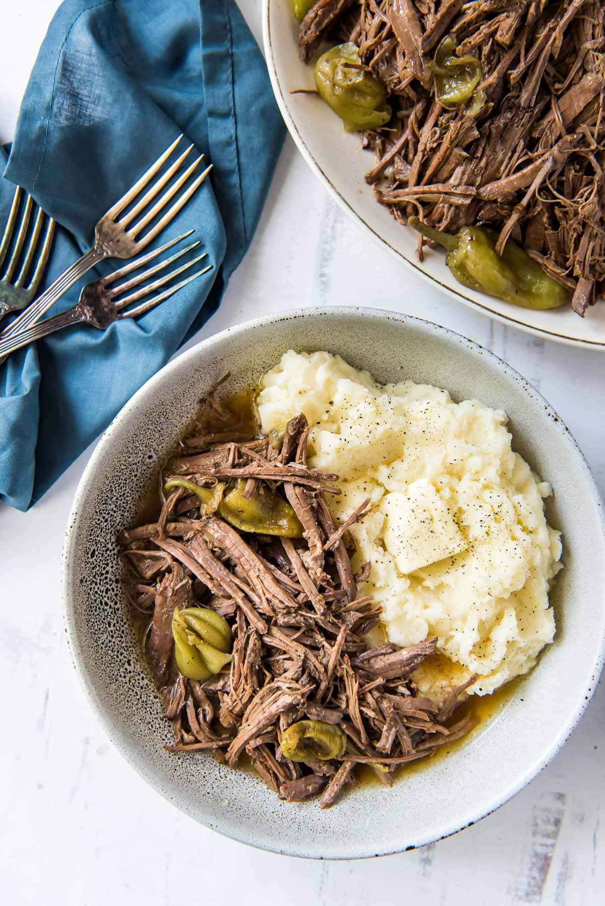 a platter with shredded top round roast, a bowl with shredded meat and mashed potatoes, a blue napkins, three forks