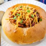 Bread bowl full of chili with a sprinkle of cheese and green onions.