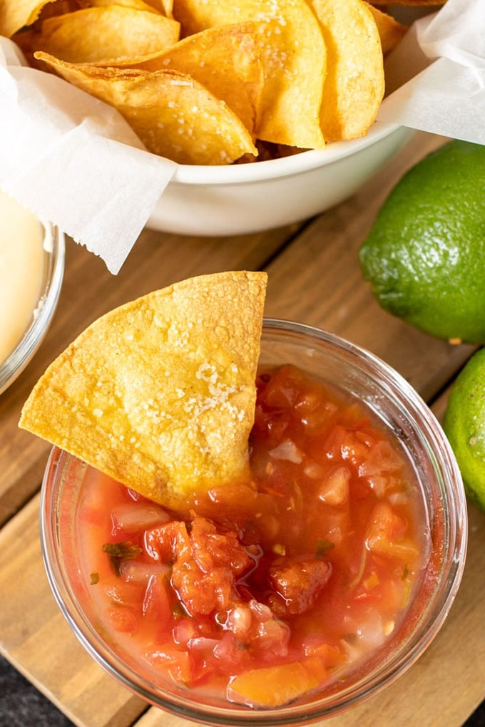 a white bowl lined with white paper and filled with tortilla chips, a clear dish of salsa with a tortilla chip in int, 2 limes