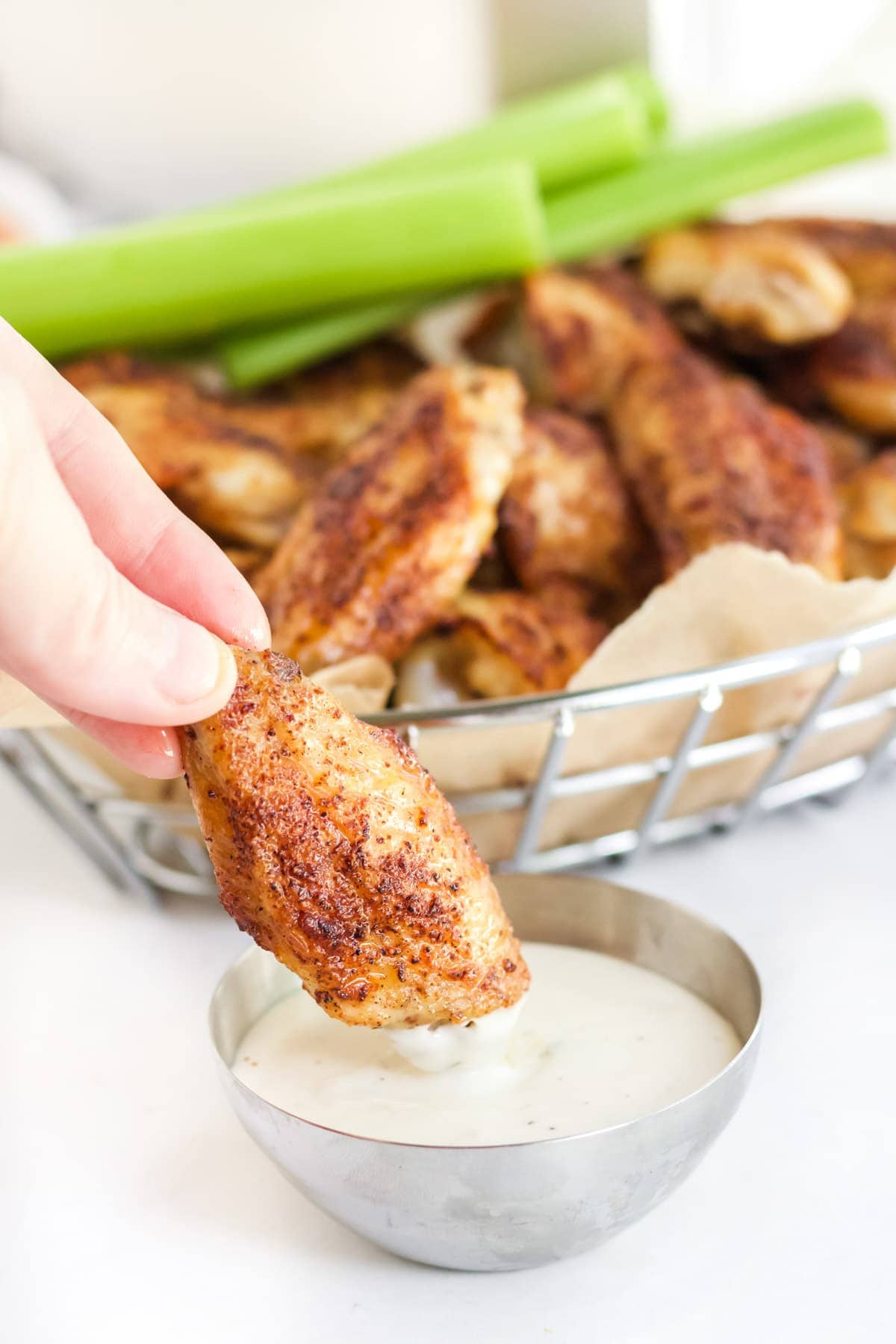 a hand, chicken wing dipped in a dish of ranch, basket of chicken wings, celery