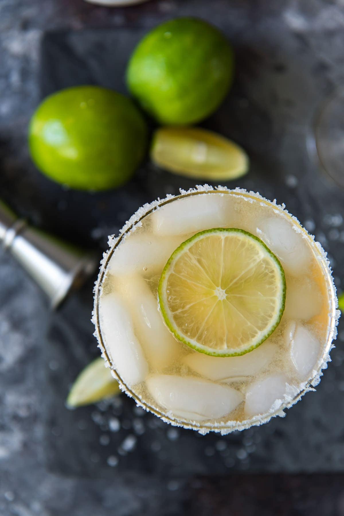 overhead shot of glass of margarita with jigger and limes below on a black background