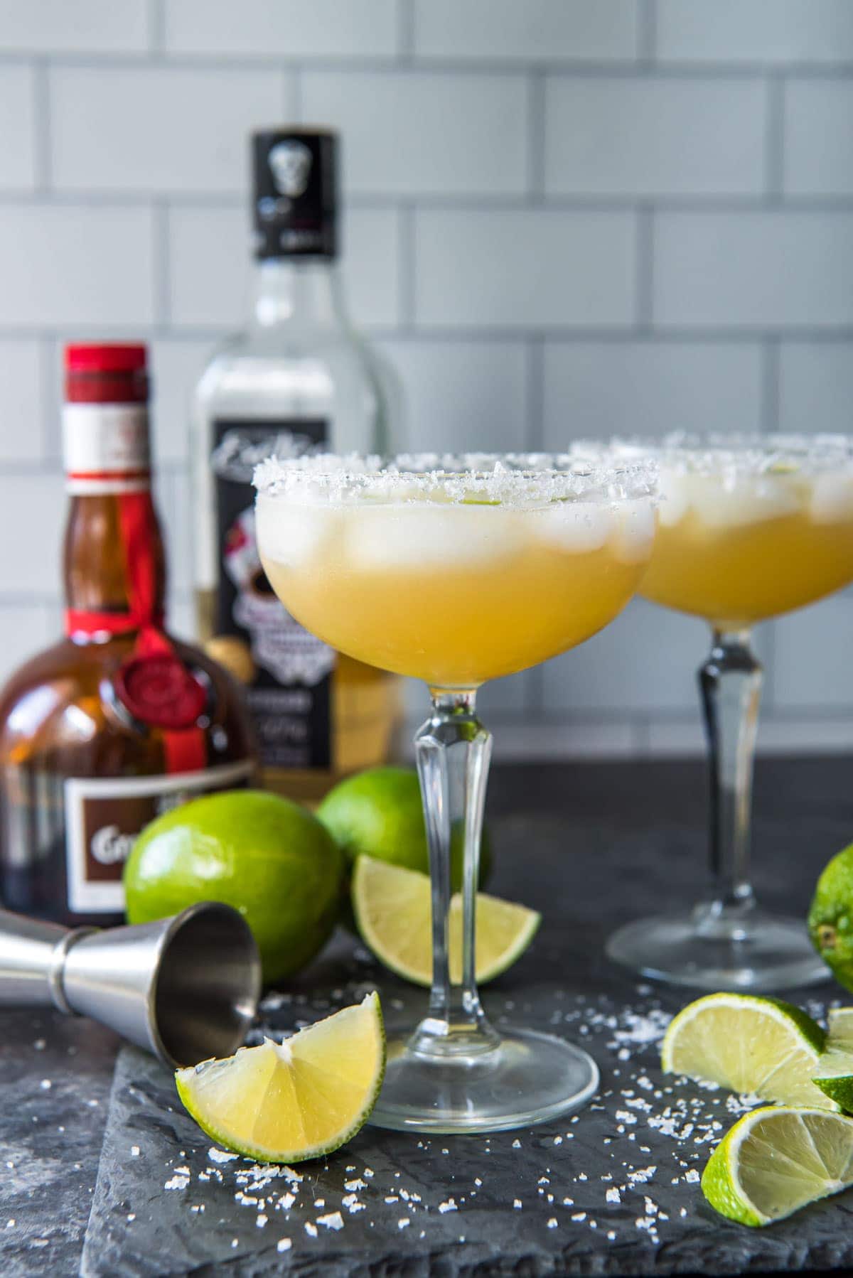 black background, lime wedges, glasses filled with cadillac margaritas, tequila, grand marnier bottles