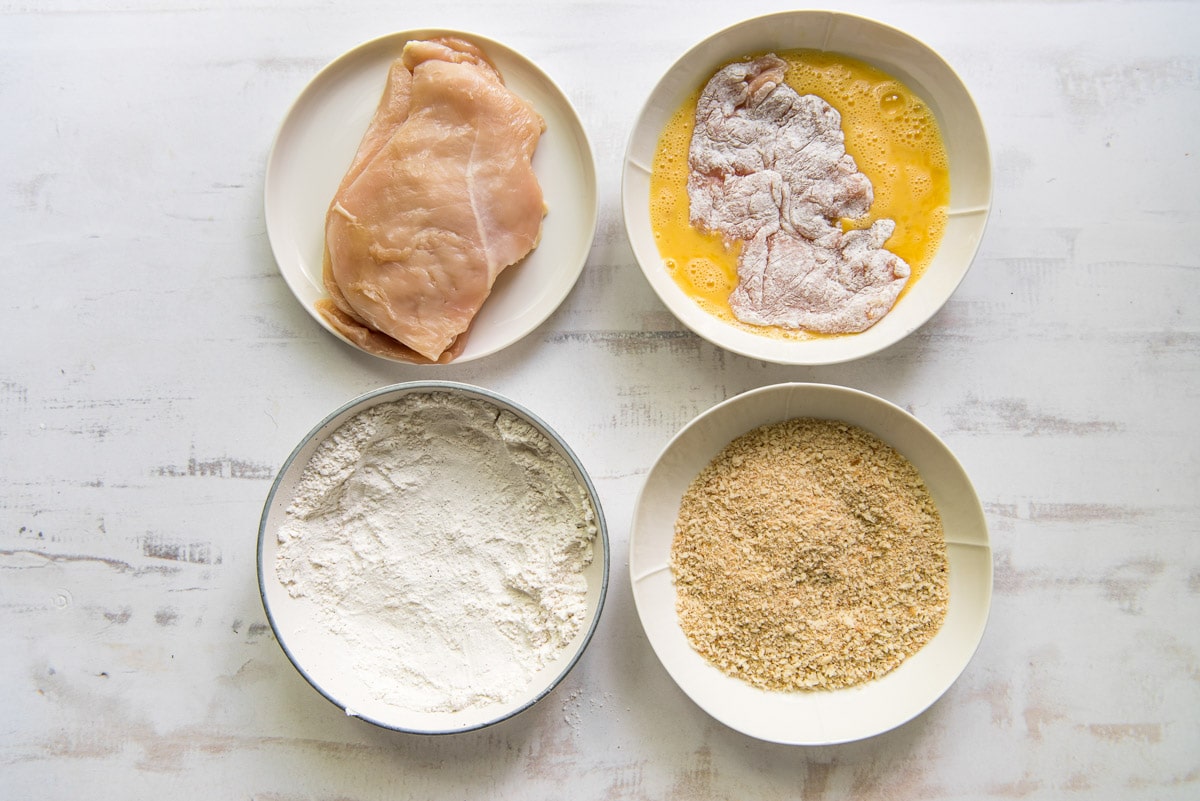 Four shallow white bowls, one with chicken cutlets, one with whisked eggs, one with flour and one with breadcrumbs.