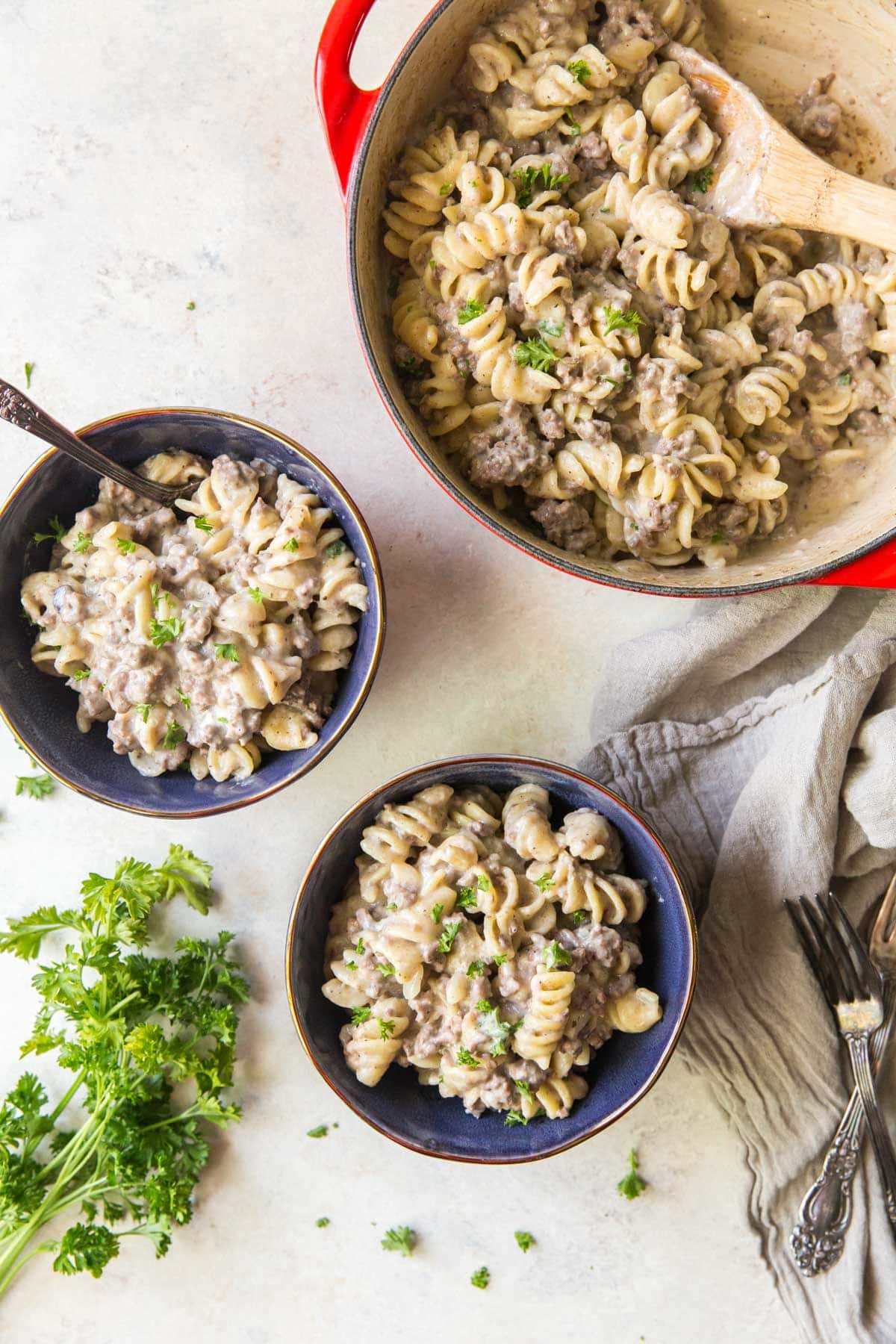 2 blue bowls with beef stroganoff, a large red pot, a wooden spoons, beige napkins, forks