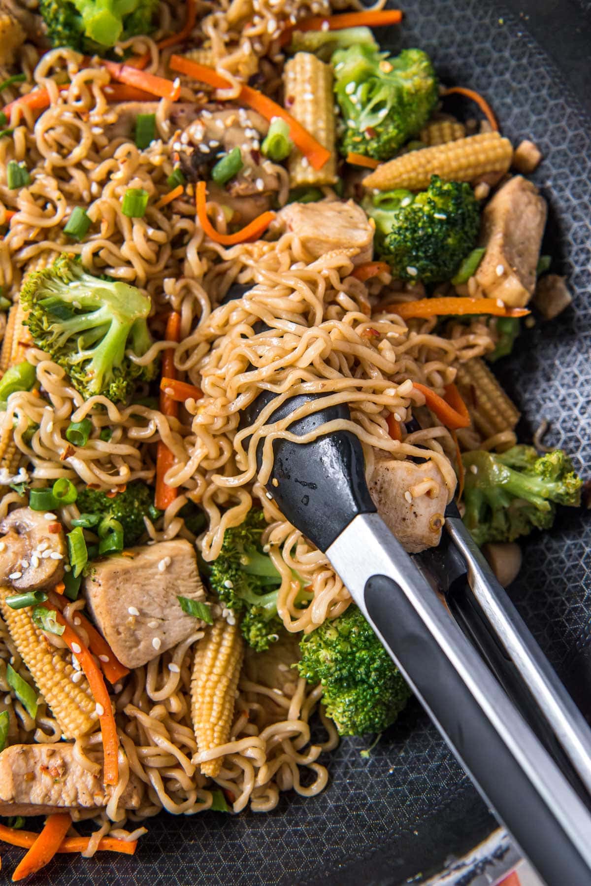 skillet with ramen noodles, stir fry with chicken and vegetables, black and silver metal tongs