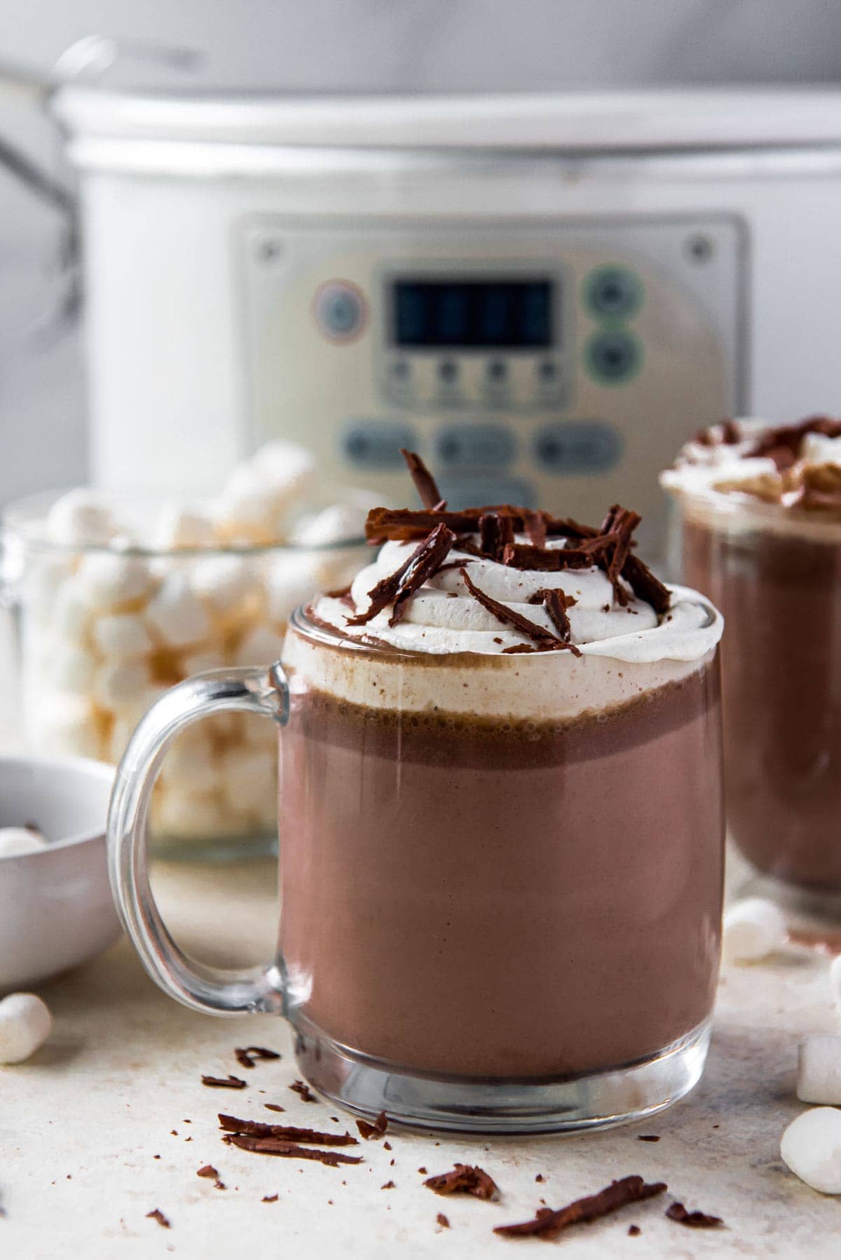 white slow cooker, hot chocolate in a clear glass, whipped cream, chocolate shavings, marshmallows