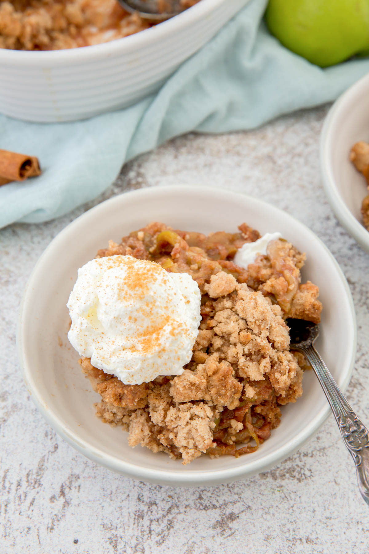 white bowl with apple cobbler, a spoons, whipped cream, cinnamon stick, blue napkin, green apple