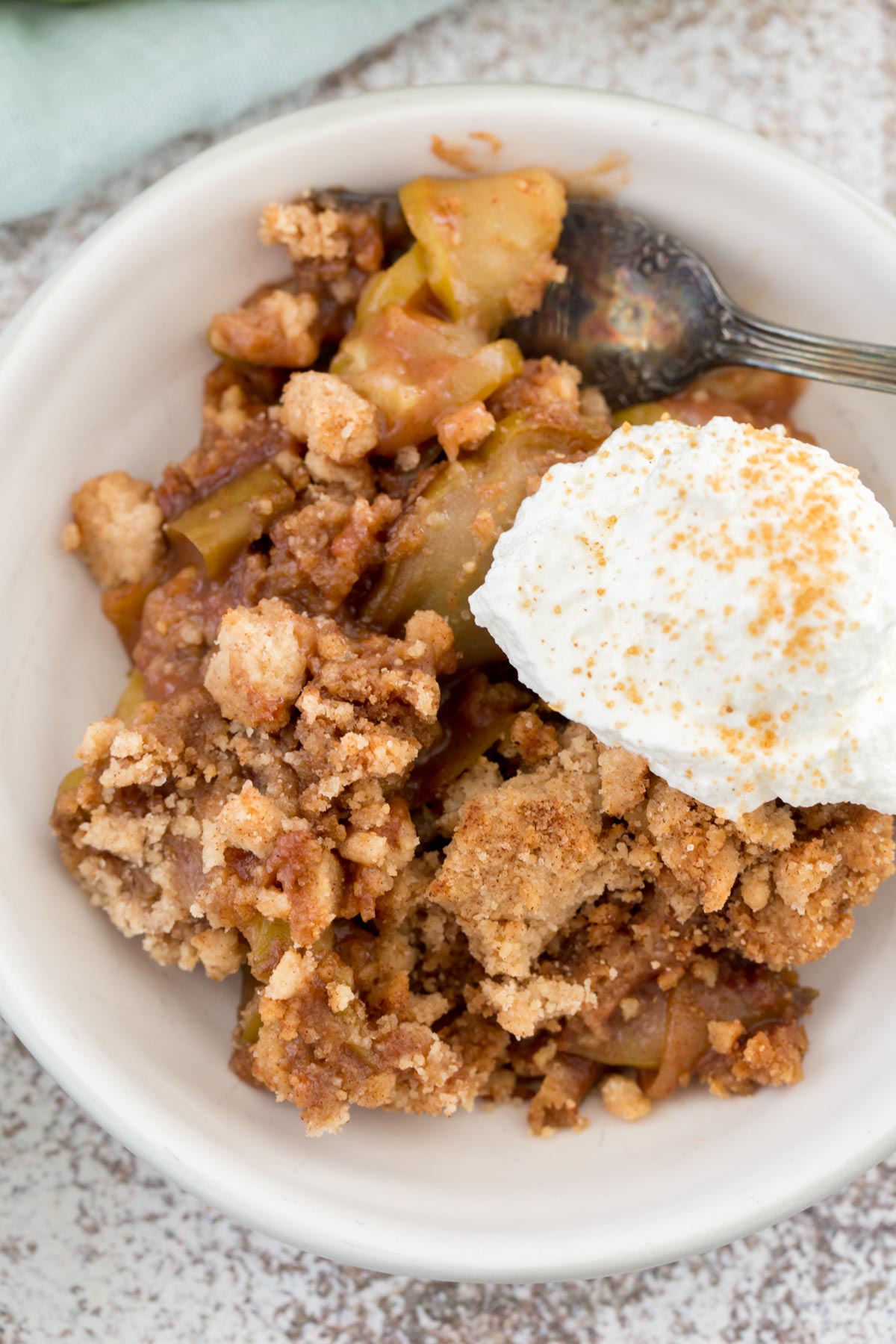 white bowl, apples, cookie topping, whipped cream, cinnamon, spoon